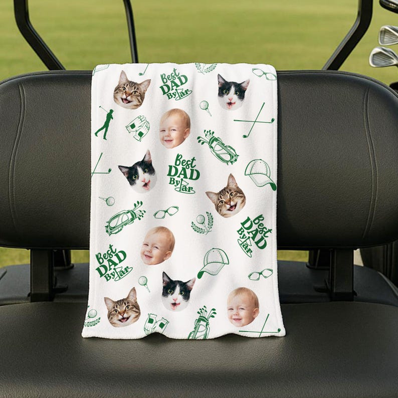 Puede incluir: Toalla de golf blanca con un patr&oacute;n repetido de caras de gatos, caras de beb&eacute;s, gr&aacute;ficos de golf y el texto "Best Dad By Far" en verde. La toalla est&aacute; colocada sobre el respaldo de un asiento de carrito de golf.