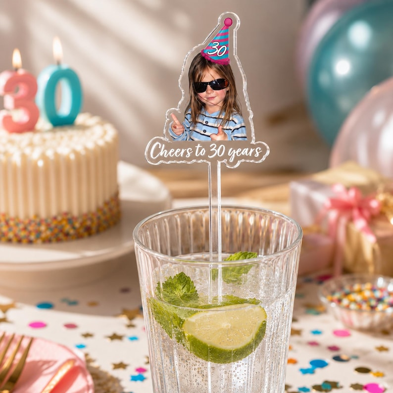 Puede incluir: Un agitador de bebidas de acr&iacute;lico transparente con un recorte de una persona con gafas de sol y un gorro de fiesta rosa y azul con el n&uacute;mero 30, con el texto Cheers to 30 years impreso en blanco en la base del dise&ntilde;o.