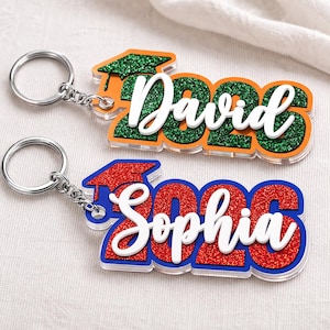 May include: Two personalized graduation keychains. One is orange with green glitter and the name "David" and the year "2023". The other is blue with red glitter and the name "Sophia" and the year "2023". Both have silver keyrings.