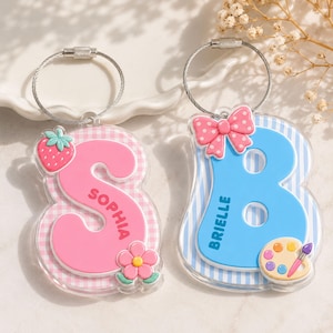 May include: Two acrylic letter keychains with metal wire loops. The pink letter S features a gingham pattern, a strawberry, a flower, and the name Sophia. The blue letter B features a striped pattern, a pink polka dot bow, and a paint palette.
