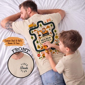 May include: Beige t-shirt with a construction-themed design on the back, featuring a road, vehicles, and the text "DAD & NOLAN CONSTRUCTION CO." The front of the shirt has the text "TIRED Dads CLUB". A child plays with toy cars near the shirt.