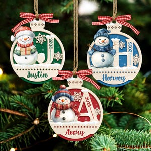 May include: Three round wooden Christmas ornaments featuring snowmen, red and white gingham bows, and twine hangers. Each ornament displays a name, Justin, Harvey, or Avery, the year 2025, and a large letter J, H, or A in green, blue, or red.