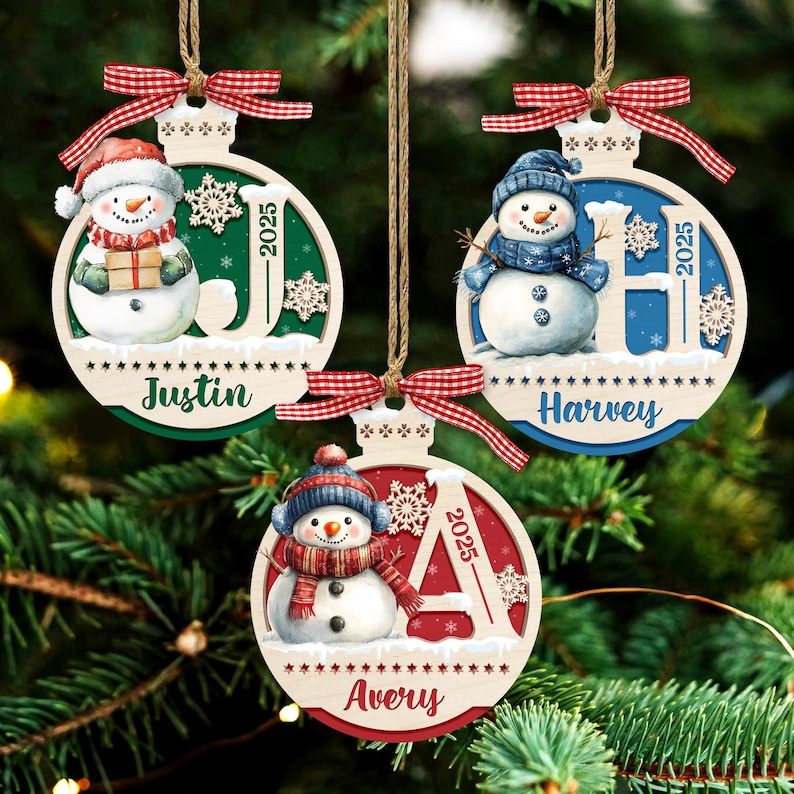 May include: Three round wooden Christmas ornaments featuring snowmen, red and white gingham bows, and twine hangers. Each ornament displays a name, Justin, Harvey, or Avery, the year 2025, and a large letter J, H, or A in green, blue, or red.