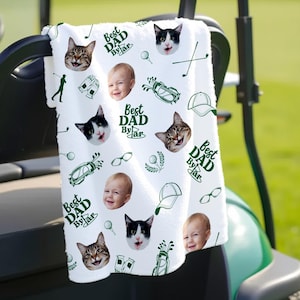 Puede incluir: Toalla de golf blanca con ilustraciones verdes con tem&aacute;tica de golf y el texto "Best Dad By Far". La toalla est&aacute; decorada con im&aacute;genes de gatos, beb&eacute;s, palos de golf, pelotas de golf y sombreros. La toalla est&aacute; colgada en un carrito de golf.