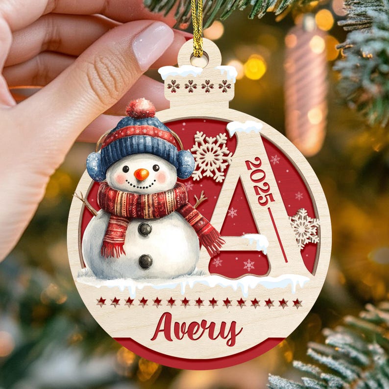 May include: A round wooden Christmas ornament featuring a snowman wearing a blue hat and red scarf, a large letter A with the year 2025, and the name Avery printed at the bottom. The design includes white snowflakes on a red background.