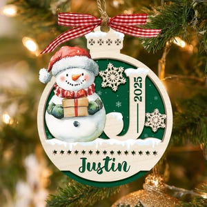 May include: A round wooden Christmas ornament featuring a snowman wearing a red hat and scarf, holding a gift. The design includes a large letter J, the year 2025, and the name Justin in green text. A red and white gingham ribbon hangs from the top.