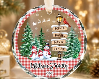 May include: A round glass Christmas ornament with a red and white gingham border. The center features a snowy forest scene with three snowmen, a street sign labeled Sophia, Daniel, Max, and Clover, a cardinal, and the text Wilson Family 2026.