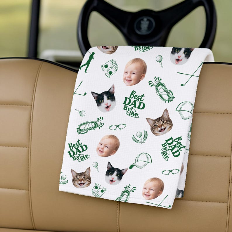 Puede incluir: Toalla de golf blanca con un patr&oacute;n repetido de caras de gatos, caras de beb&eacute;s, gr&aacute;ficos de golf y el texto "Best Dad By Far" en verde. La toalla est&aacute; doblada sobre el asiento de un carrito de golf.