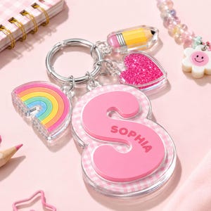 May include: A silver metal keychain featuring a large, clear acrylic letter S with a pink gingham background and the name SOPHIA printed in pink. Attached are three smaller acrylic charms: a colorful rainbow, a pink glitter heart, and a yellow pencil.