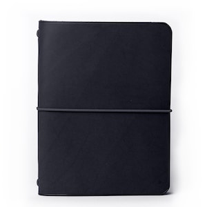 Op de afbeelding: Een rechthoekige zwarte notitieboekomslag met afgeronde hoeken, voorzien van een gladde matte textuur en een enkele horizontale elastische band die om het midden is gewikkeld om de omslag gesloten te houden tegen een witte achtergrond.