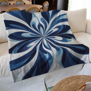 Puede incluir: Una manta blanca con un patrón floral abstracto azul y blanco. La manta está doblada y colocada sobre un sofá blanco.