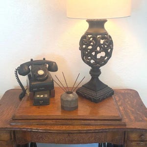 Puede incluir: Un teléfono de disco vintage negro con cable en espiral, un pequeño difusor de cerámica gris con varillas de madera y una lámpara de mesa negra con base calada intrincada y pantalla blanca, todo sobre una superficie de madera.