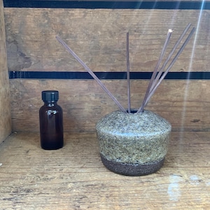 Könnte beinhalten: Eine kleine, dunkelbraune Glasflasche mit schwarzem Deckel steht neben einer gesprenkelten Keramikvase. Die Vase enthält mehrere Räucherstäbchen. Die Vase hat ein hellgrünes und braunes Farbschema. Die Gegenstände stehen auf einer Holzoberfläche.