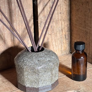 Może przedstawiać: Teksturowany dyfuzor z kamienia z brązowymi patyczkami i małą butelką z brązowego szkła z czarną zakrętką. Dyfuzor ma sześciokątną podstawę i cylindryczny korpus. Przedmioty znajdują się na drewnianej powierzchni.