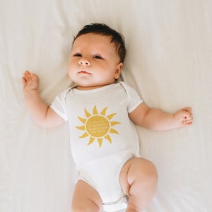 Puede incluir: Un bebé con un body blanco con un gráfico de sol amarillo y el texto "Rayo de sol pequeño, iluminando nuestros días".