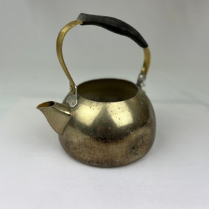 Può includere: Un bollitore d'argento con manico nero e dettagli dorati. Il bollitore ha un corpo arrotondato e un beccuccio laterale.