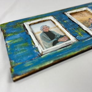 Op de afbeelding: Een rustiek houten fotolijst met vier openingen, geschilderd in tinten blauw en groen. Elke opening heeft een wit frame en toont een andere foto.