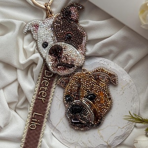 May include: A beaded keyring featuring two dog heads, one white and brown and one golden brown, attached to a gold heart-shaped clasp. A mauve velvet ribbon with the embroidered text "Dieter Lilo" in gold thread hangs from the top dog head.