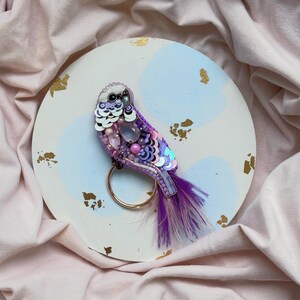 Op de afbeelding: Een broche in de vorm van een vogel in paars en roze met iriserende pailletten en kralen. De broche is op een witte achtergrond met blauwe en gouden accenten.