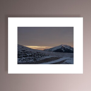 May include: A snowy mountain peak with a hint of pink in the sky at sunset. The mountain is in the distance with a field of snow and grass in the foreground.