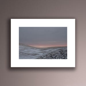 May include: A snowy landscape with a pink and purple sunset in the distance. The snow-covered hills are rolling and the sky is a soft, muted color.