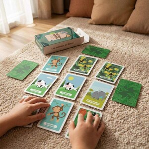 Peut inclure: Un jeu de cartes intitulé Les animaux du Coeur comprenant une boîte verte et plusieurs cartes rectangulaires illustrées d'animaux comme un singe, une vache, un éléphant et des abeilles, avec du texte français, disposées sur une surface texturée.