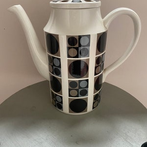 May include: A white ceramic coffee pot with a geometric pattern of black, brown and grey circles and squares. The pot has a lid with a white knob.
