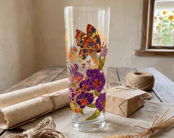 May include: A clear glass vase featuring hand-painted designs of a vibrant orange and yellow butterfly, purple flowers with green leaves, and a small bird. The artwork is rendered in a stained glass style with defined outlines on the cylindrical surface.