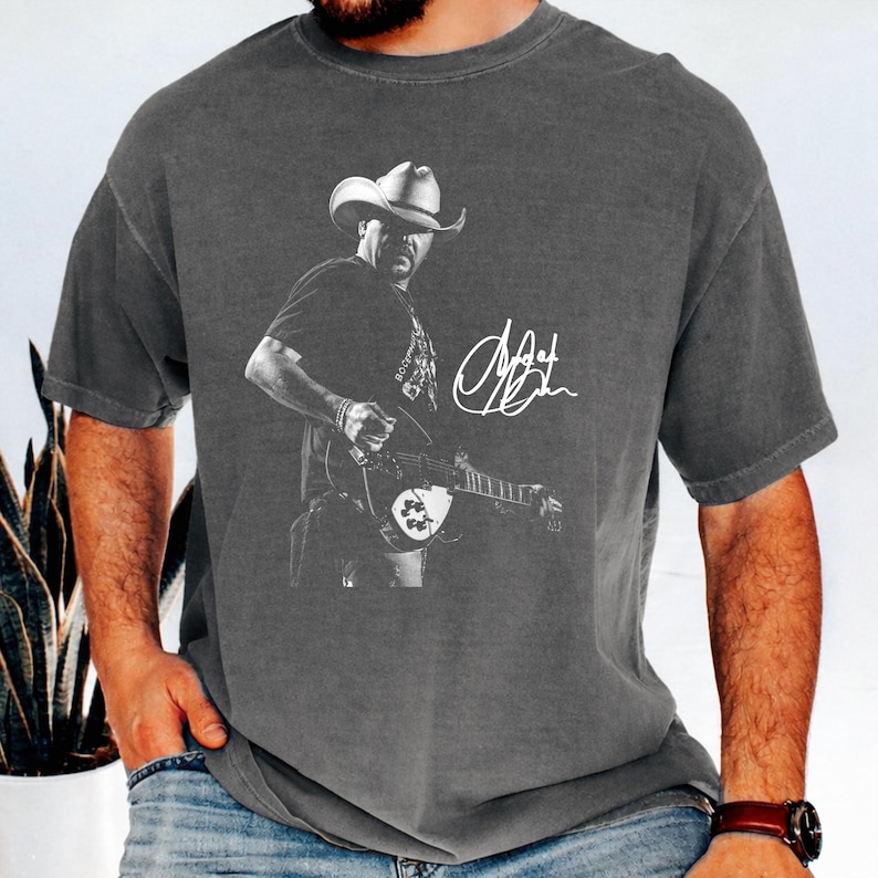 May include: A charcoal gray short-sleeve t-shirt featuring a black and white graphic of a musician wearing a cowboy hat and playing an electric guitar. The design includes a white handwritten-style signature graphic positioned to the right side.