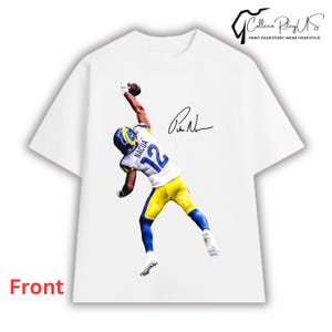May include: A white short-sleeve t-shirt featuring a graphic of a football player in a white, blue, and yellow uniform with the number 12 and the name NACUA on the back, reaching up to catch a football, accompanied by a black signature.