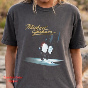 May include: A charcoal gray short-sleeve t-shirt featuring the gold cursive text Michael Jackson above a graphic of black shoes with white socks standing on a reflective surface. The shirt has a crew neckline and a relaxed fit design.
