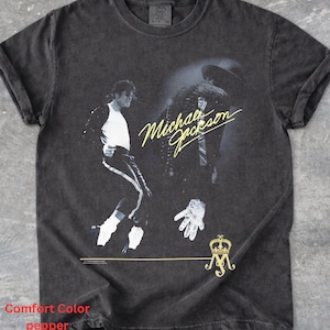 May include: A charcoal grey short-sleeve t-shirt featuring a graphic print of Michael Jackson in a dance pose, a large white glove, and the text Michael Jackson in yellow script. A gold crown and MJ monogram logo are printed at the bottom right.
