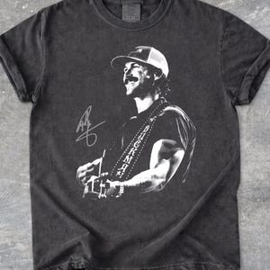 May include: A charcoal gray short-sleeve t-shirt featuring a white graphic print of a musician wearing a baseball cap and playing a guitar. The guitar strap displays the text DUCKMAN, and a signature is printed on the left side of the graphic.