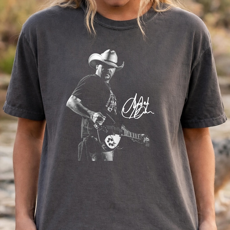 May include: A charcoal gray short-sleeve t-shirt featuring a white graphic print of a musician wearing a cowboy hat and playing an electric guitar. The design includes a stylized white signature next to the musician, centered on the chest.