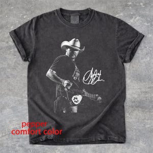 May include: A pepper-colored t-shirt featuring a black and white graphic of a musician wearing a cowboy hat and playing an electric guitar. A white signature is printed to the right of the musician. The shirt has a crew neck and short sleeves.