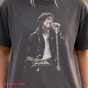 May include: A charcoal gray short-sleeve t-shirt featuring a black and white graphic print of Michael Jackson singing into a microphone. The design includes the name Michael Jackson in a cursive script font below the image. The shirt is labeled Comfort Color pepper.