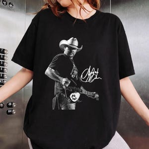 May include: A black short-sleeve t-shirt featuring a monochromatic graphic print of a musician wearing a cowboy hat and playing an electric guitar. The design includes a white handwritten-style signature positioned to the right of the figure.