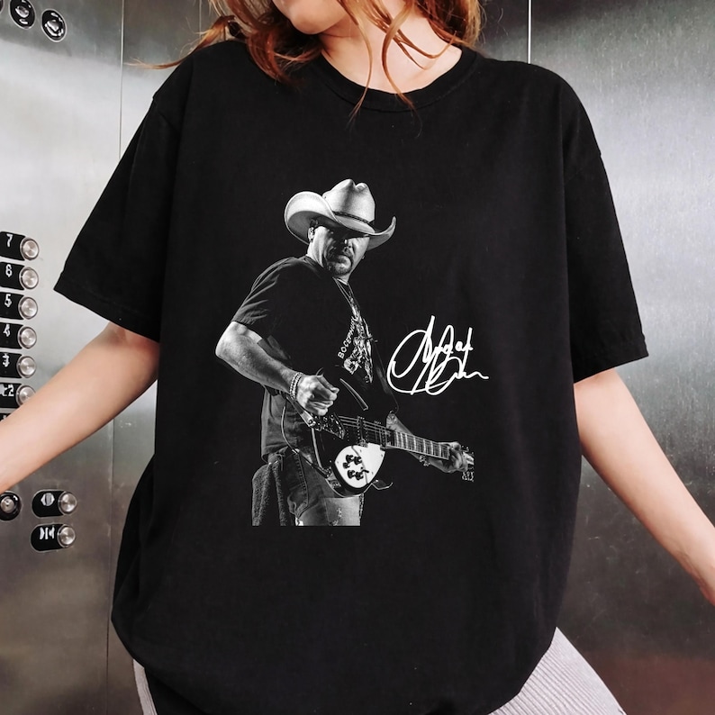 May include: A black short-sleeve t-shirt featuring a monochromatic graphic print of a musician wearing a cowboy hat and playing an electric guitar. The design includes a white handwritten-style signature positioned to the right of the figure.