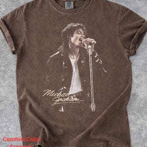 May include: A brown short-sleeve t-shirt featuring a graphic print of Michael Jackson singing into a microphone. The shirt has a crew neckline and a faded, vintage-style finish. The text "Michael Jackson" is printed in a script font below the image.
