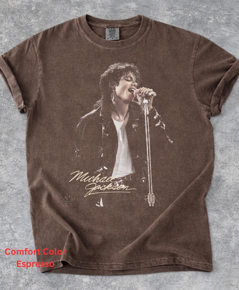 May include: A brown short-sleeve t-shirt featuring a graphic print of Michael Jackson singing into a microphone. The shirt has a crew neckline and a faded, vintage-style finish. The text "Michael Jackson" is printed in a script font below the image.