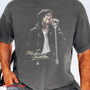 May include: A charcoal gray short-sleeve t-shirt featuring a black and white graphic print of Michael Jackson singing into a microphone. The shirt has a vintage-style washed appearance and includes the name Michael Jackson in white script text.