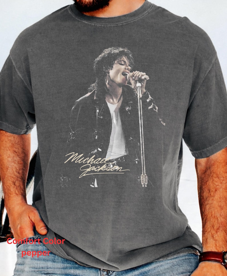 May include: A charcoal gray short-sleeve t-shirt featuring a black and white graphic print of Michael Jackson singing into a microphone. The shirt has a vintage-style washed appearance and includes the name Michael Jackson in white script text.