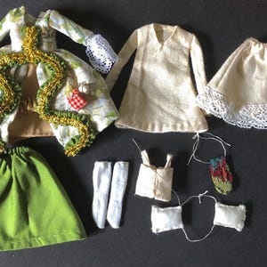 May include: A collection of doll clothing including a floral patterned coat with gold trim and white lace cuffs, a solid green skirt, a cream-colored long-sleeve tunic, a cream skirt with lace trim, white stockings, a small vest, and accessories.