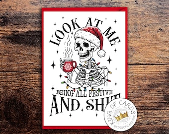May include: A rectangular greeting card featuring a black and white illustration of a skeleton wearing a red Santa hat, holding a red mug with a snowflake design, and draped in colorful Christmas lights, with the text LOOK AT ME BEING ALL FESTIVE AND SHIT.
