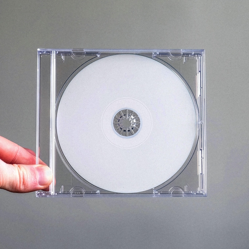 May include: A clear plastic CD jewel case containing a blank white compact disc. The disc features a standard circular silver center hub with a textured gear-like pattern. The case is transparent, showcasing the plain white surface of the disc.