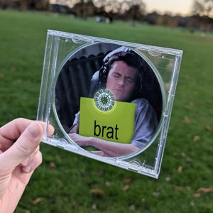 May include: A clear plastic CD jewel case containing a disc with a printed image of a person wearing headphones and a grey shirt, holding a bright lime green square with the word brat printed in black lowercase letters on the front.