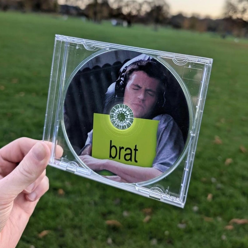 May include: A clear plastic CD jewel case containing a disc with a printed image of a person wearing headphones and a grey shirt, holding a bright lime green square with the word brat printed in black lowercase letters on the front.