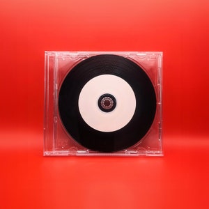 Può includere: Una custodia per CD in plastica trasparente con un CD nero all'interno. Il CD ha un'etichetta bianca con un piccolo design circolare argentato al centro.