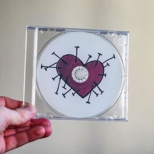Puede incluir: Un CD en una funda de plástico transparente. El CD tiene un fondo blanco con un diseño de corazón granate atravesado por alfileres negros. El corazón está centrado en el disco.