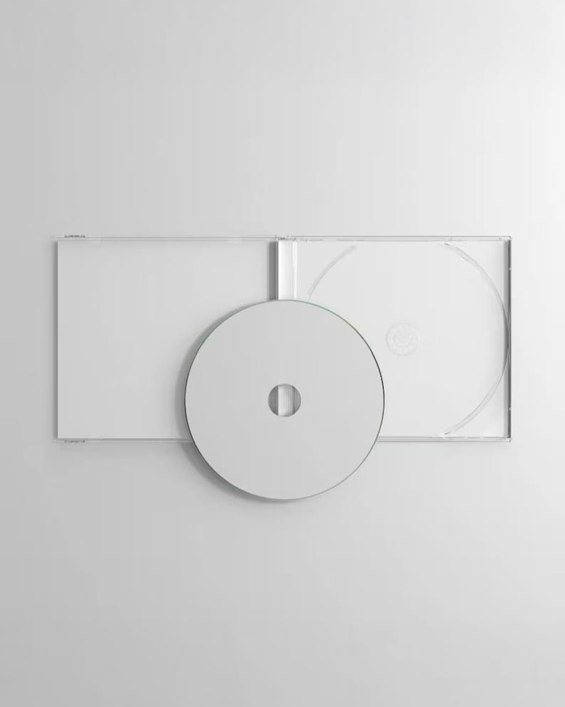 May include: A clear plastic CD jewel case is open and laid flat, revealing the interior tray. A blank, solid white circular compact disc is positioned in front of the open case, partially obscuring the center of the clear plastic housing.